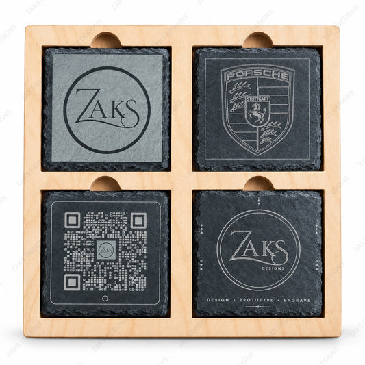 A set of four square dark slate coasters arranged in a light natural wood tray with four compartments and circular cutouts. The coasters feature engraved designs including a circular 'Zaks' logo, a Porsche crest, a QR code, and the words 'Design - Prototype - Engraved'.