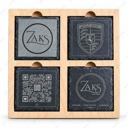 A set of four square dark slate coasters arranged in a light natural wood tray with four compartments and circular cutouts. The coasters feature engraved designs including a circular 'Zaks' logo, a Porsche crest, a QR code, and the words 'Design - Prototype - Engraved'.